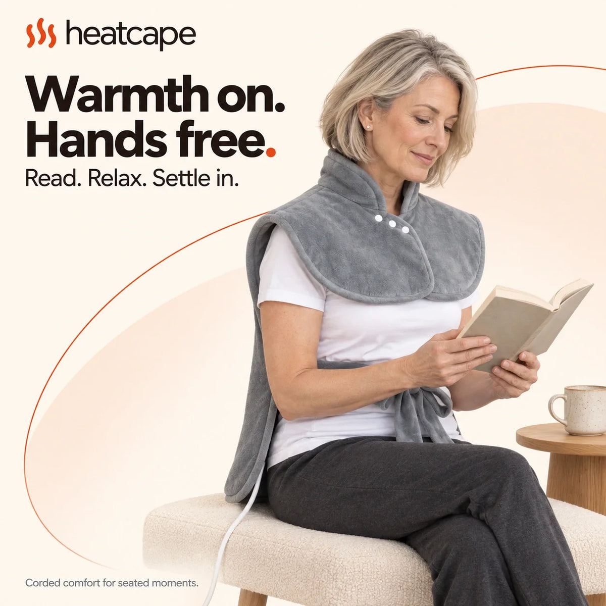 A man reading in an armchair while wearing the Warmith wrap, its cable visible beside the chair.