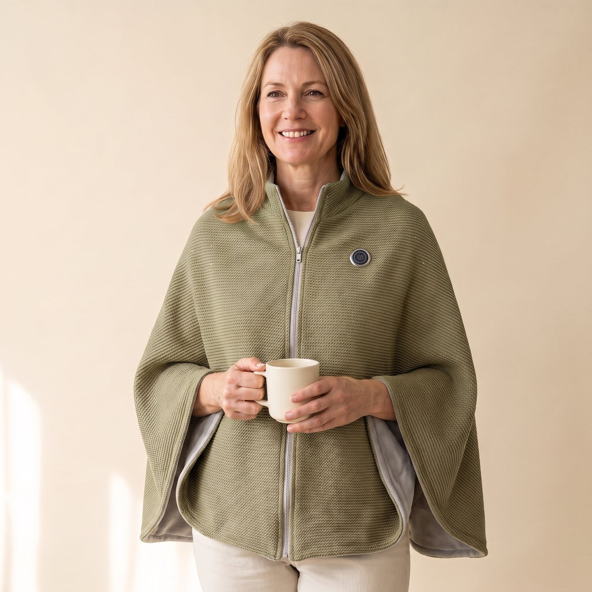 Woman wearing the olive Warmith heated shawl wrap zipped, holding a mug